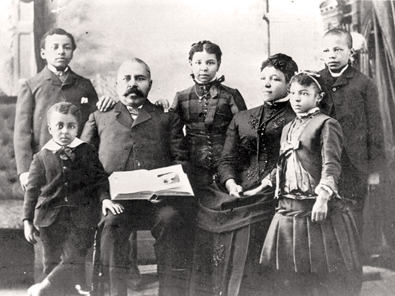 Richard and his wife, America sit in chairs surrounded by their 5 children who are standing.