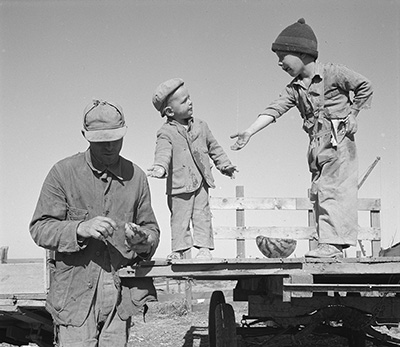 Two young boys stand on a wood platform about waist high. A man stands on the ground next to them holding a piece of watermellon