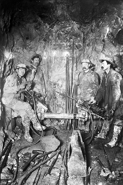 4 men covered in filth and dust pose for a photo in 1906. Each wears a hat, boots and overcoat with sturdy pants and shirt.