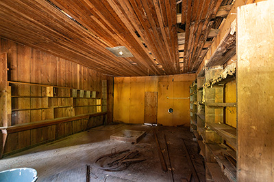 Room with wood planky floor, ceiling and walls with empty shelves. All is falling apart & dirty with dust and cobwebs.