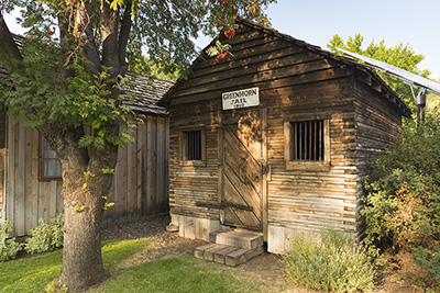 Wooden 1 room building with 2 windows covered in bars on front wall. Sign says "Greenhorn Jail 1910"