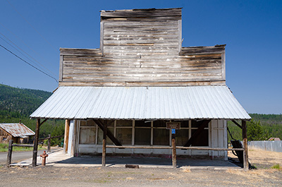 Wood plank building with false facade. Sheet metal roof covers the front wood walkway. Surrounded by gravel, dirt & dead grasses