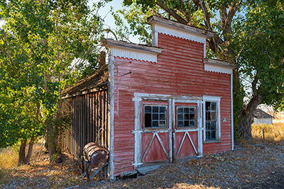 An old wood building with a facade extending above the true dimensions. Trees grow close around it.