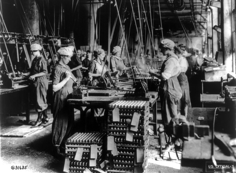 Photo of women in work clothes stand at benches full of machine parts.