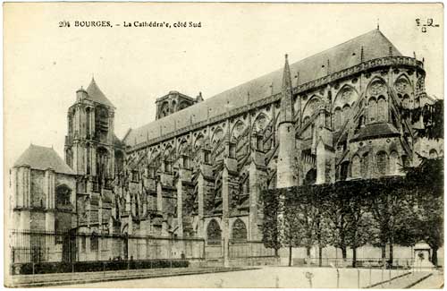 Cathedral with many spires, two towers, a fence and short trees in front.