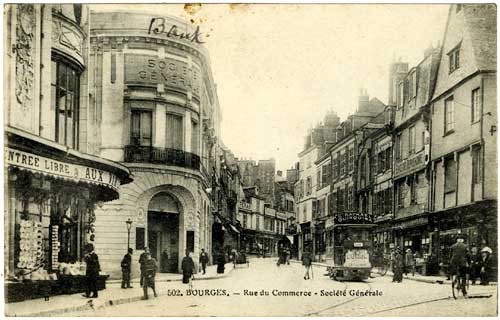 Business lined street, Bourges France, tall buildings each side, a street car on track down the right, people walking and biking