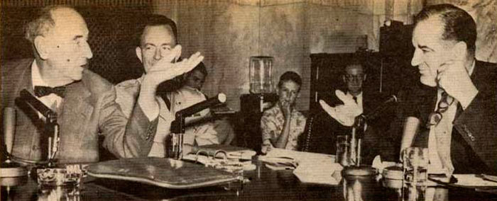 Photo of General Joseph Welch talking across a table to Senator McCarthy. There are microphones, papers & binders on the desk.