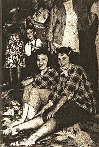 Photo of 2 young women in matching paid shirts sit on the floor or on a low bench at a party.