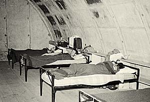 3 men, each lies on a cot, inside a cement igloo.