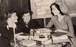 Katharine Hepburn sits next to Chico Marx and talks to a woman sitting to the other side of Chico. The table in front has books.