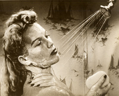 A woman stands under a shower head and holds a bar of soap in her hand.