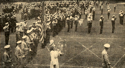 Crowd of people in parade formation, some carrying flags, some insturments. All wearing hats and military finery.