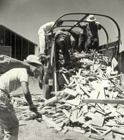 Men stand in the back of a truck filled with scrap lumber pushing it off the end of the truck bed.