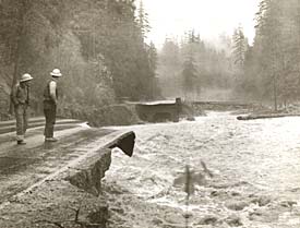 2 men stand at the edge of a washed out road with flood waters going by.