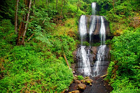 a waterfall falls over 3 rock terrace formations. Lush green foliage grows on the banks at the sides.