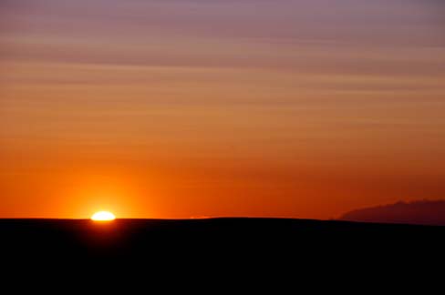 half of sun showing up over ridge of landscape. Orange streaks in sky.