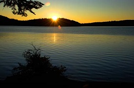 Calm lake with banks cloaked in darkness the sun peeks our from behind a hill as it sets.