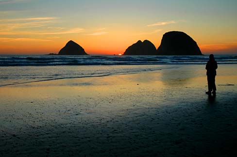 Person standing on beach at sunset with 3 rockformations out in ocean.