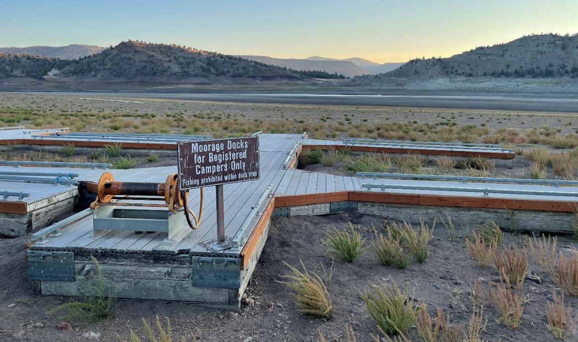 A dock sits on dry ground in front of a lake with very low water