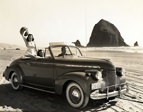 A man driving a car with a woman in the passenger seat & another woman sitting high in the back. The woman in back holds a ball.