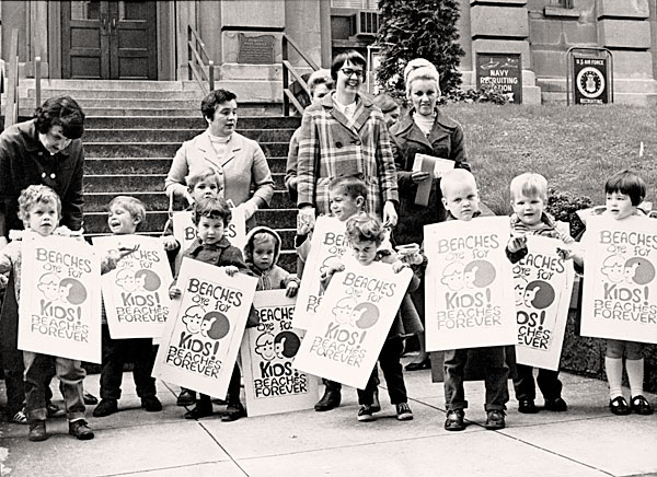 5 women stand behind 10 children stand in front of a building holding signs that read: Beaches are for Kids! Beaches Forever. 