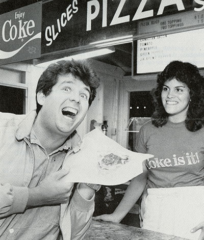 A man holds a piece of pizza a a woman watches