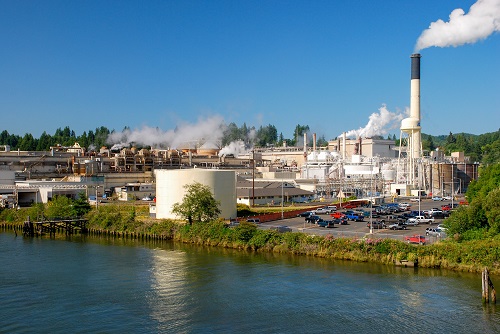 Industrial buildiings on the bank of a river. A parking lot about half fulll of vehicles is in the front.