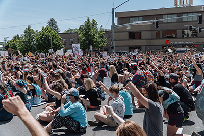 Hundreds of people sit on a paved surface all facing one way, many with their arms raised in protest.