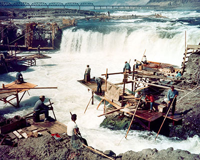 Native American men fish off platforms overhanging waterfalls.