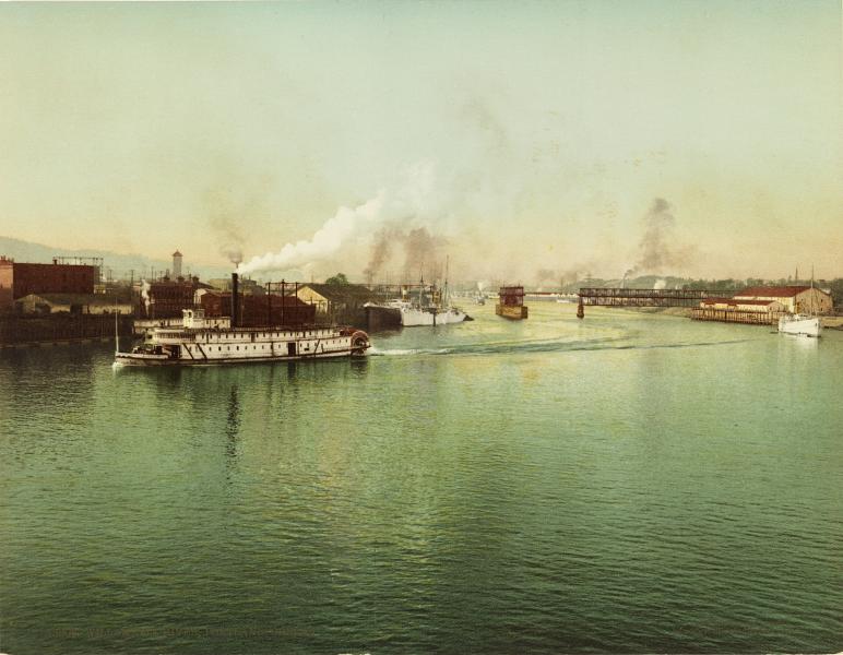 A sternwheeler boat on a river. Industrial buildings on the banks of the river.