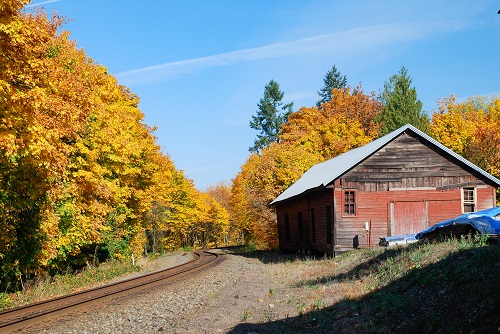 An old barn sits next to a railroad track leading to the distance. Deciduous trees line the sides in full fall color.