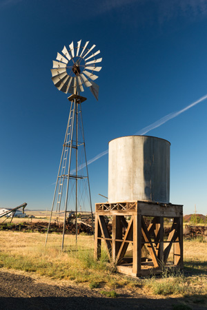 A metal windmill and metal water tank set in a grassy area next to a gravel road.