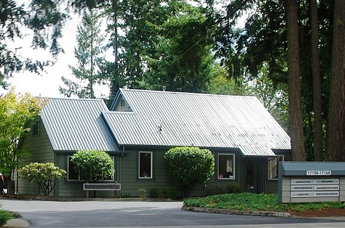 Durham City hall is an office building with a metal roof. Tall fir trees surround it and the paved parking lot.