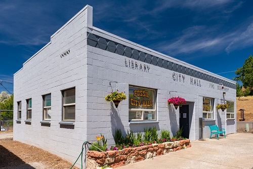 A single story cement block building serving as library and city hall in Fossil. 3 planters of flowers hang on 1 outside wall.