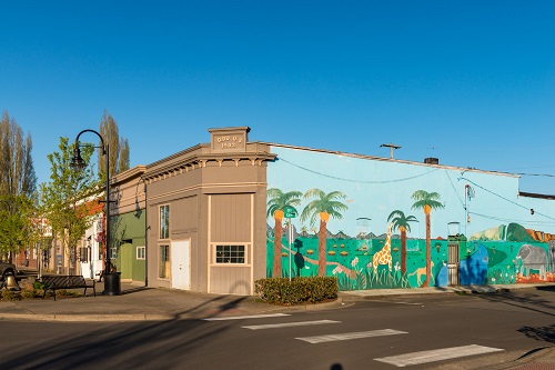 A mural on the side of a building shows palm trees, a giraffe, an elephant and other animals on a green landscape.