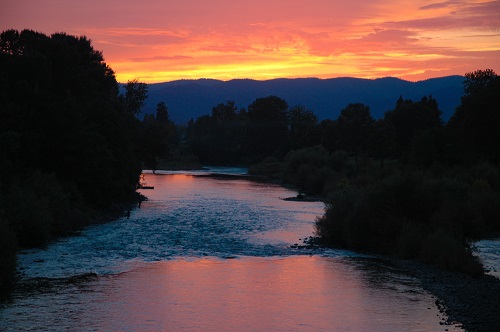 Below, a swiftly running river. On the horizon a sunset in vibrant colors. Trees line the landscape.