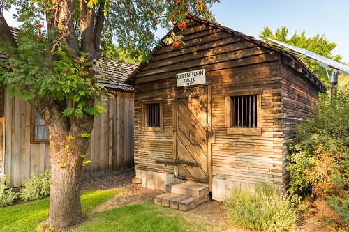 A small, square building with 2 windows in front with bars across. Thick walls of interlocking planks make the jail stout.