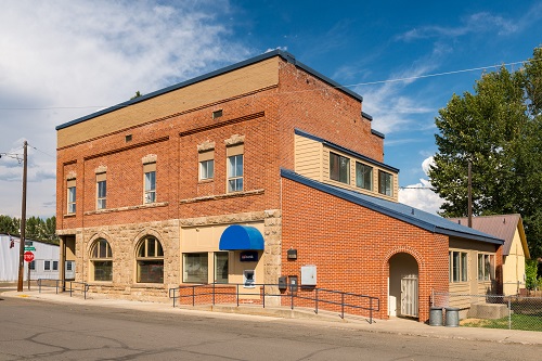 Two-story brick building takes up most of a block. 2 windows on ground level have arched tops. Windows on 2nd level are tall.