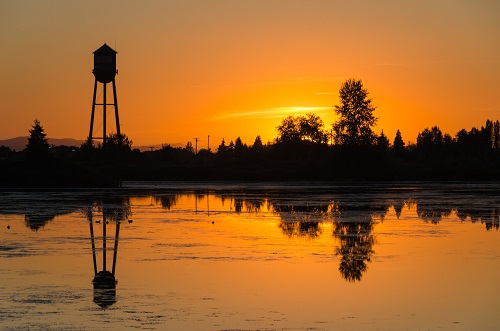 Sunset over a lake. A water tower is silhouetted on the left and trees along the bank.