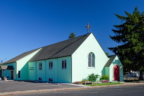 2-story church topped with a crucifix on the roof. Stain glass window, rectangle with rounded top on end of building.