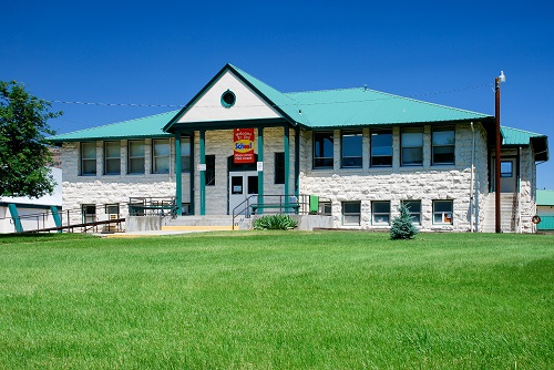 Two-story stone building with 1st level slightly sunken from ground level. Cement steps lead to double door entrance.