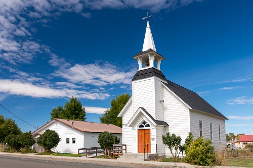 Single story church with double doors with bell tower above. 3 tall windows seen along one outside wall.