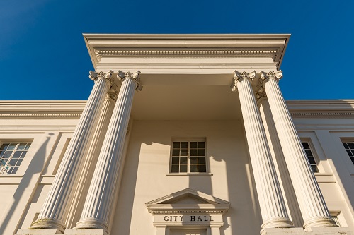 Front of city hall building with 2 Roman columns on each side of front door reaching up to 2nd story roof.