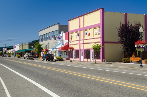 Two lane road in downtown Reedsport with runs along businesses. Baskets of summer flowers hang from lamp posts.