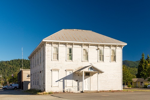 Two-story building. Wall facing us has front door with overhang covering entry. 5 tall windows on 2nd floor.