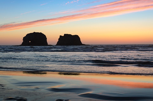 Sunset along the beach throws pink & orange colors into the sky & reflects on wet sand. Twin rock formations in the ocean near.