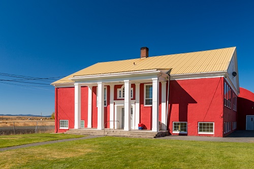 Two-story school house with double front doors. An roof overhangs the front supported by 4 columns reaching to the top.