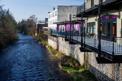 A creek runs along a stone retaining wall. On the top of the wall are businesses & a walkway with railing to look over creek.