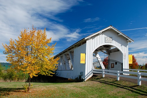 120 foot long wood covered bridge with a low wood fence lining the road leading into the bridge. 