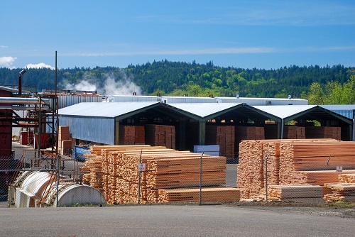 Dozens of stacks of lumber stacked on paved area. Some uncovered, some under roofs.